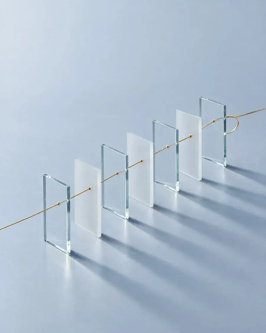 A row of glass panes on a pale grey surface, with one gold thread passing through each pane in turn